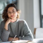 Modern ofis ortamında, not defteri ve kalem tutan kendinden emin genç profesyonel, doğal ışık ve kitaplarla çevrili.