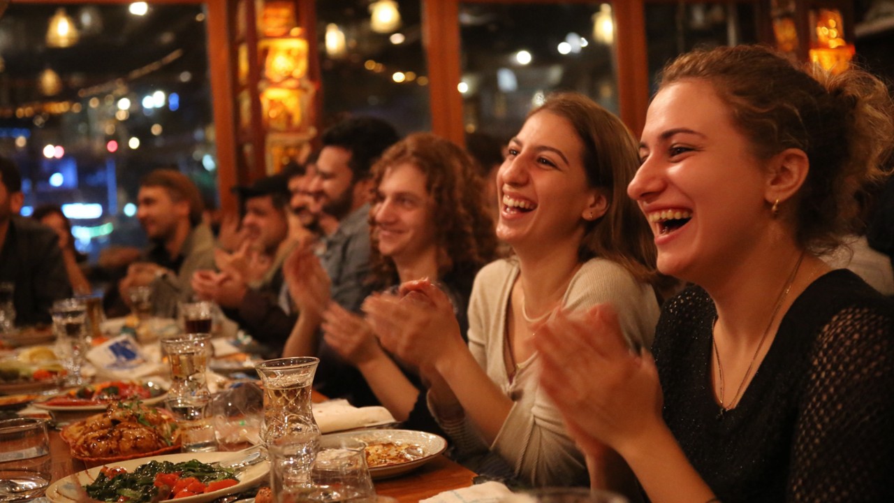 Canlı Türk rakı sofrasında gülümseyen ve alkışlayan insanlar, geleneksel yemekler ve rakı eşliğinde neşeli gece atmosferi